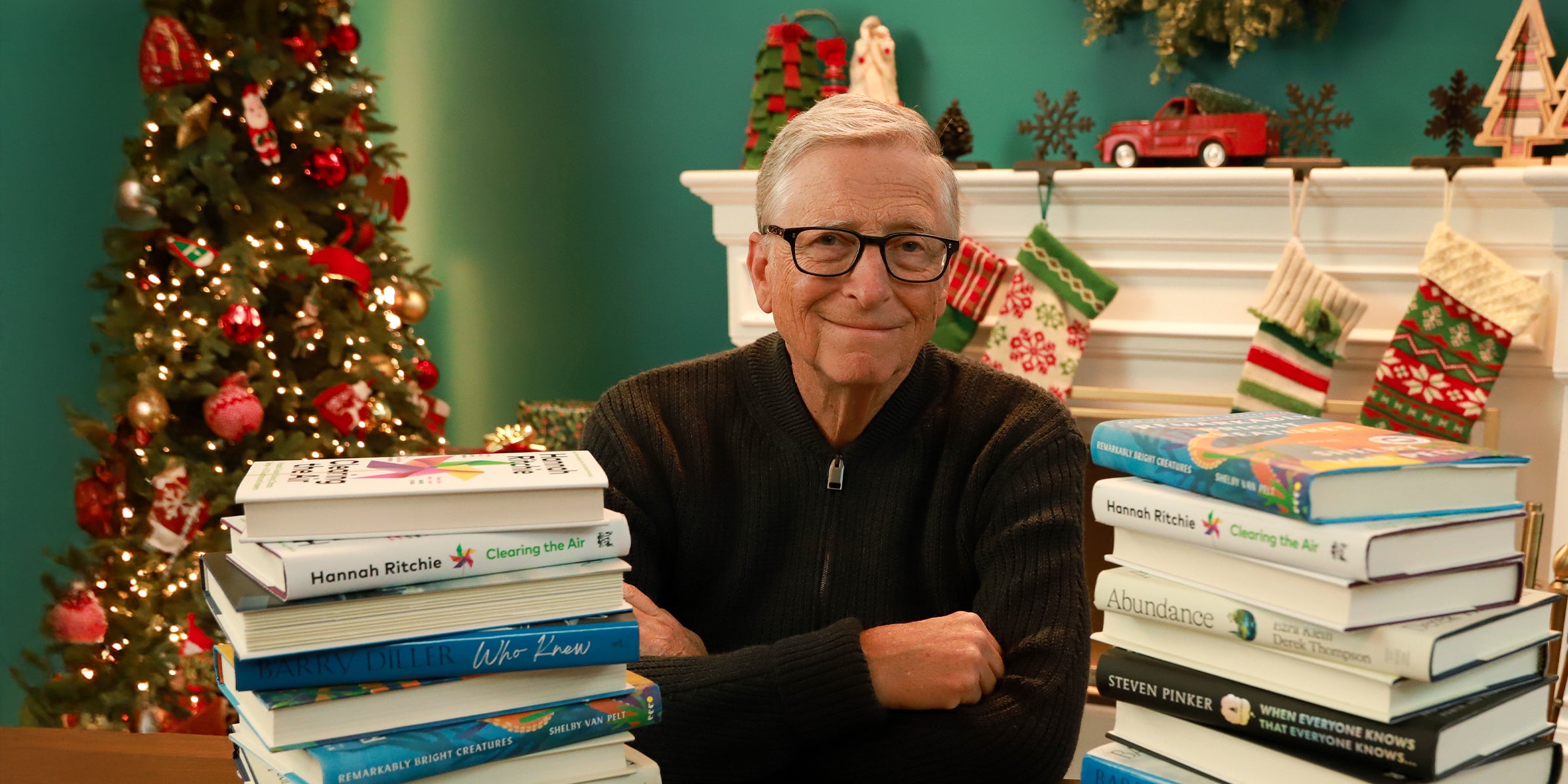 Bill Gates sitting at a table surrounded by holiday décor and stacks of books.