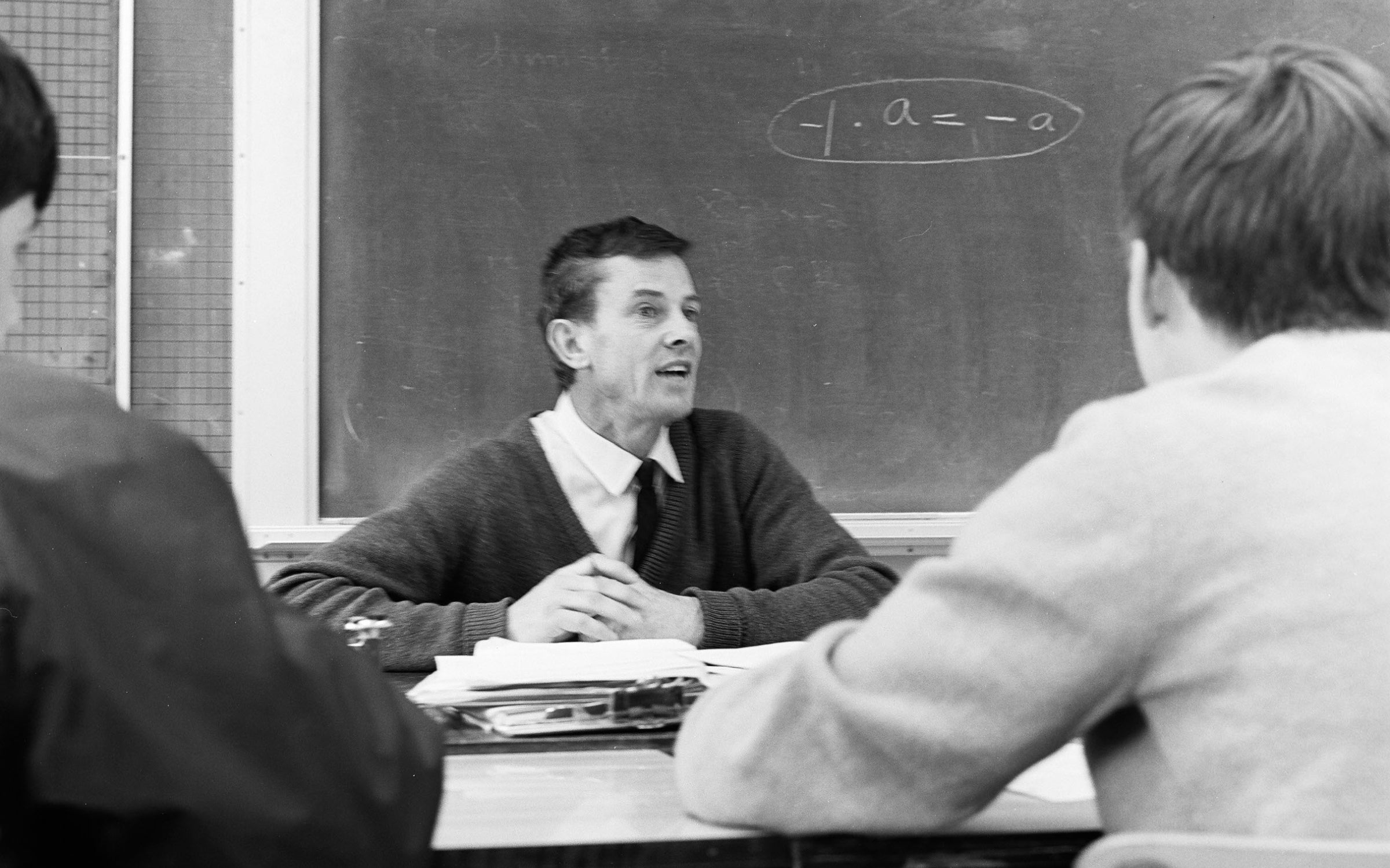Bill Dougall sitting at a desk with a blackboard in the background and students in the foreground.