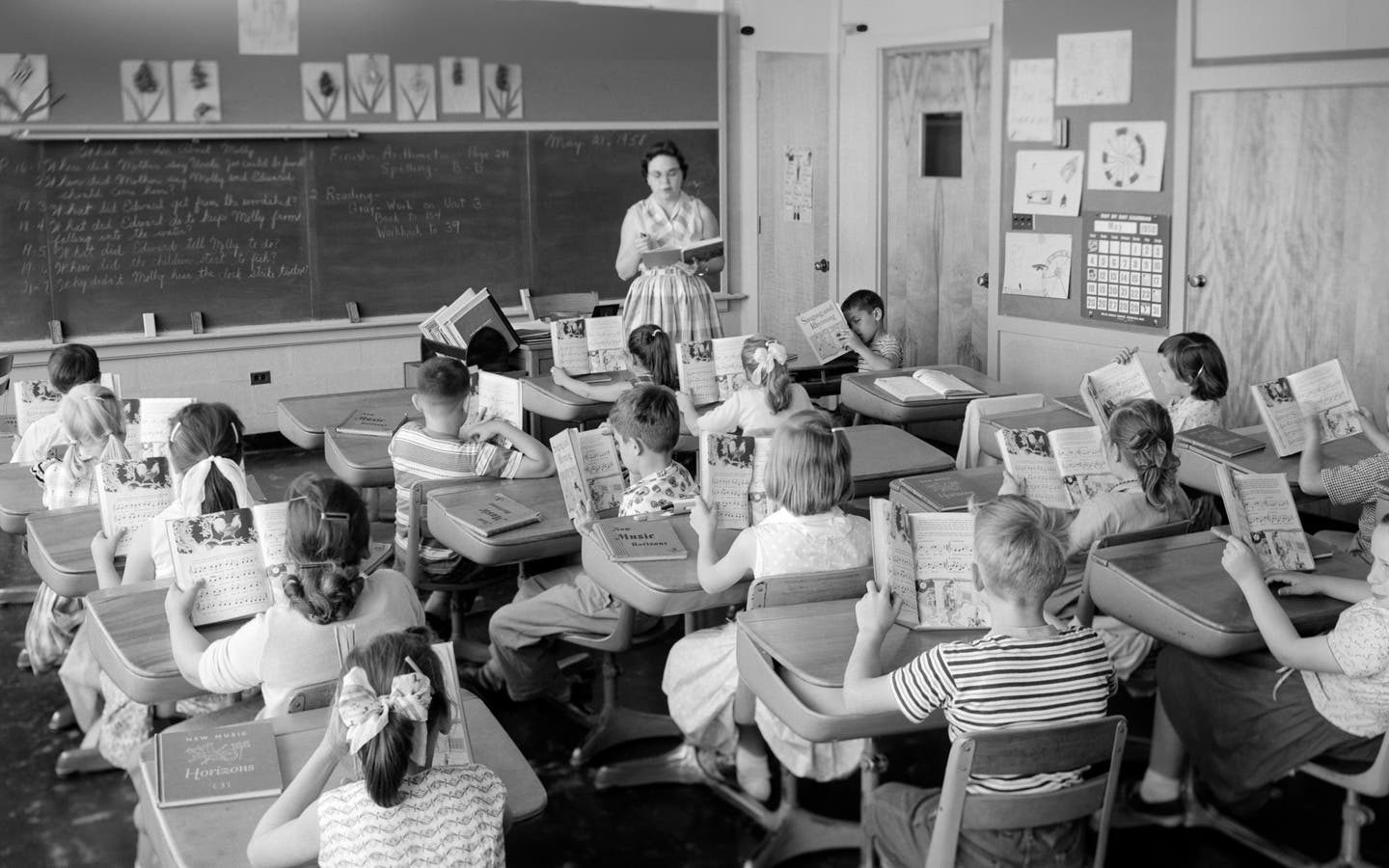 Elementary school classroom from the 1950s with a teacher standing in front of students sitting at desks reading from open textbooks.