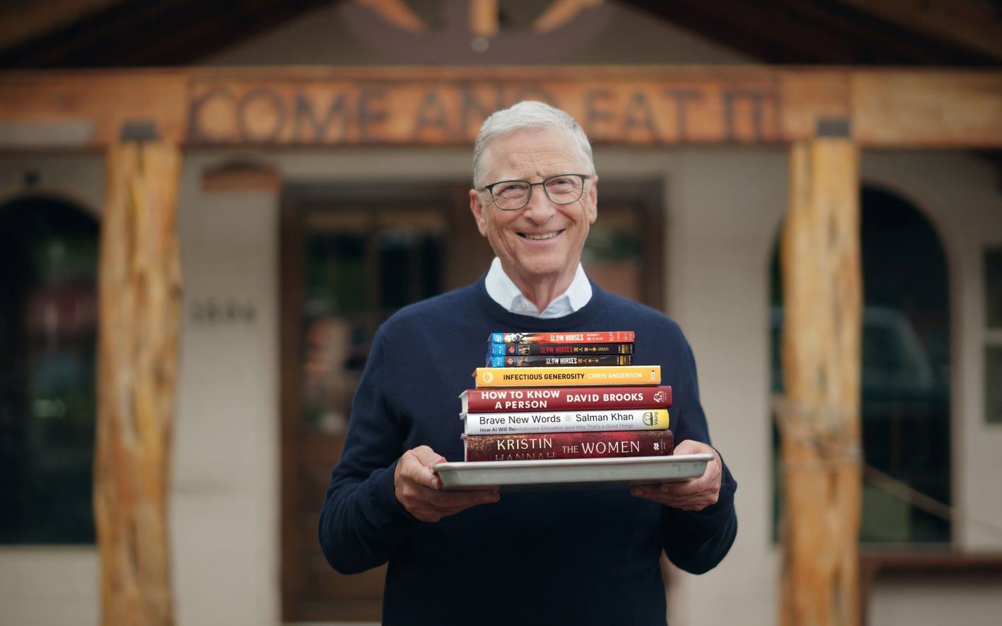 Bill Gates standing with a stack of books on a tray in front of a “come and eat” sign.