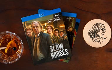 A stack of “Slow Horses” DVDs on a wood bar with an old fashioned drink and an illustration of Gary Oldman on a coaster.