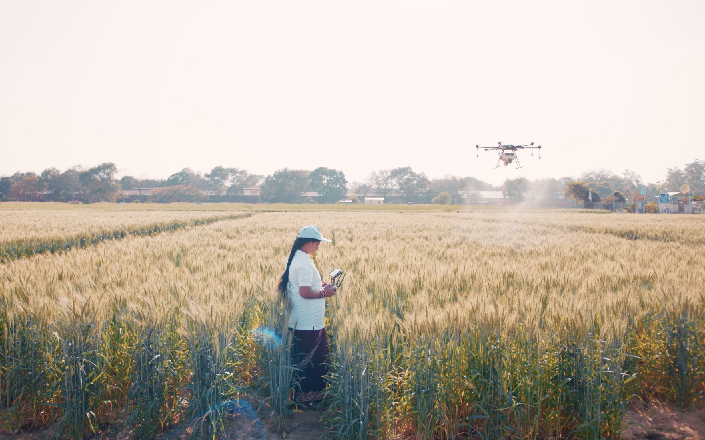 A Drone Didis standing in a field holding a drone controller device with a drone flying above spraying fertilizer.